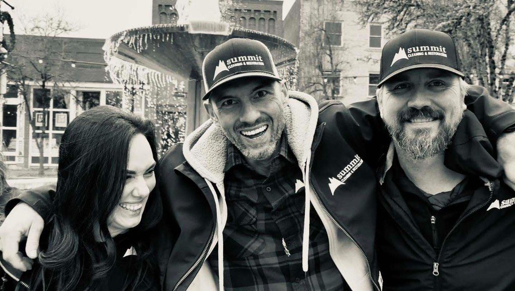 Three Summit Cleaning & Restoration team members in branded caps and jackets, arms around each other and laughing in front of a downtown fountain