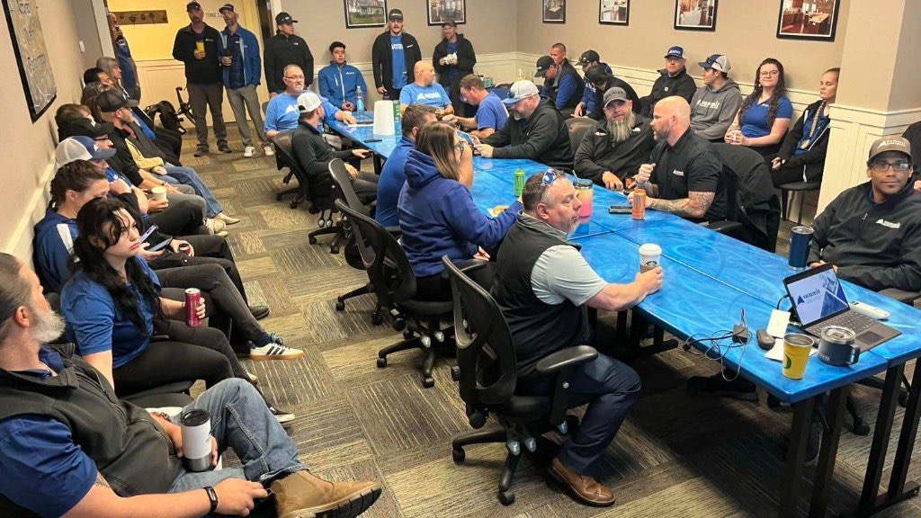 About thirty Summit employees in branded apparel seated around blue conference tables during an all-team meeting