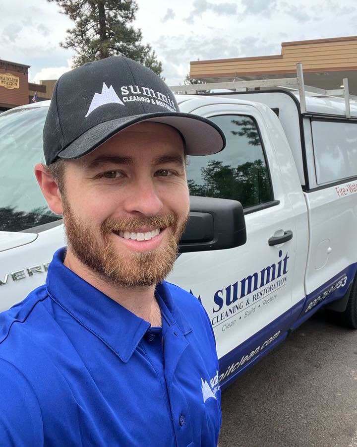 Smiling Summit technician in a blue polo and branded cap standing beside a Summit Cleaning & Restoration service truck