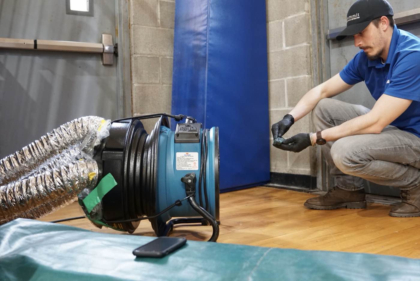 Summit technician Jordan Zeller in a branded cap and polo, crouched beside a blower connected to flexible ducting on a job site